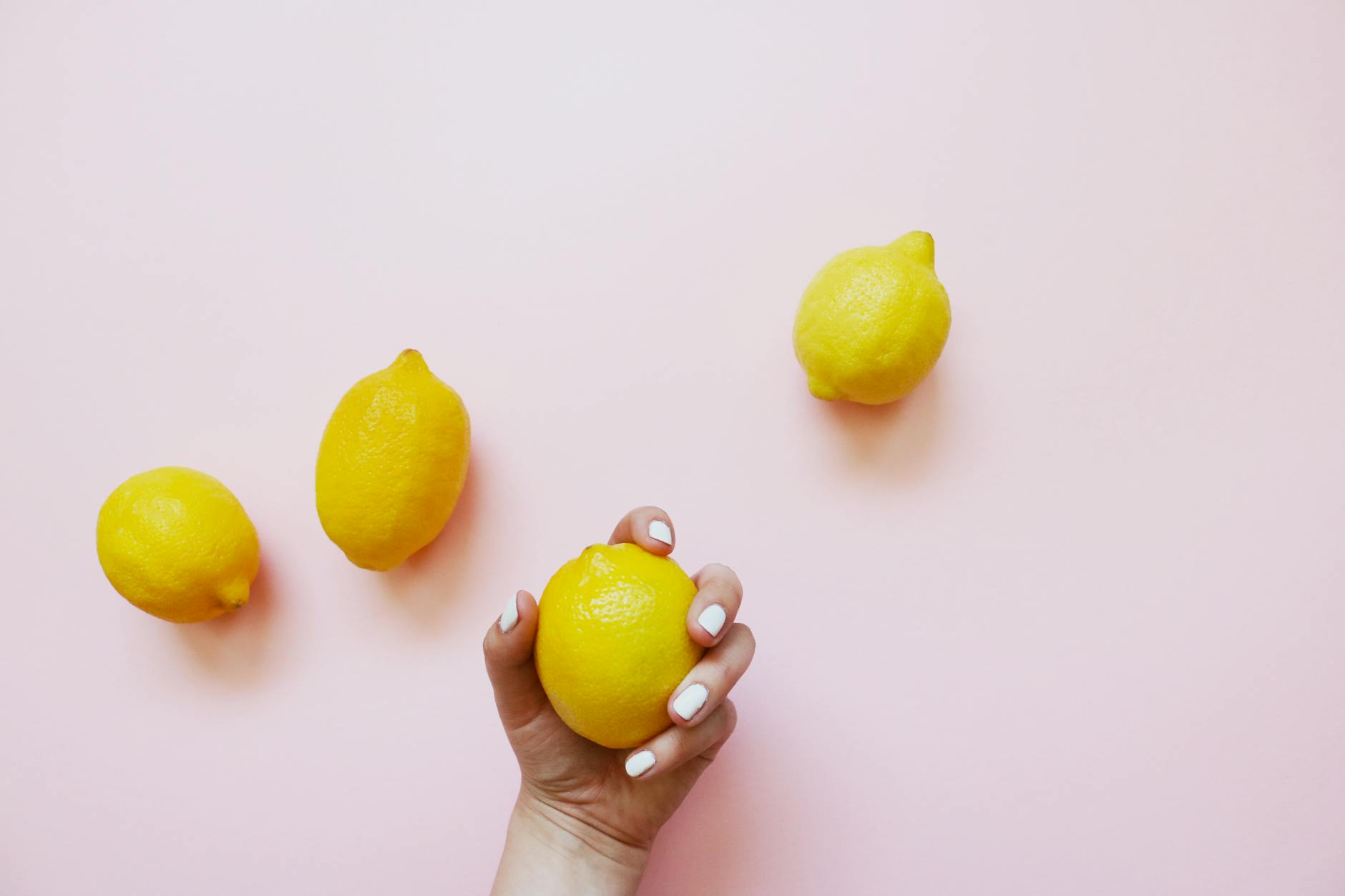 Een hand met witte nagels houdt een citroenvrucht vast op een zachte roze achtergrond, omringd door drie extra citroenen.