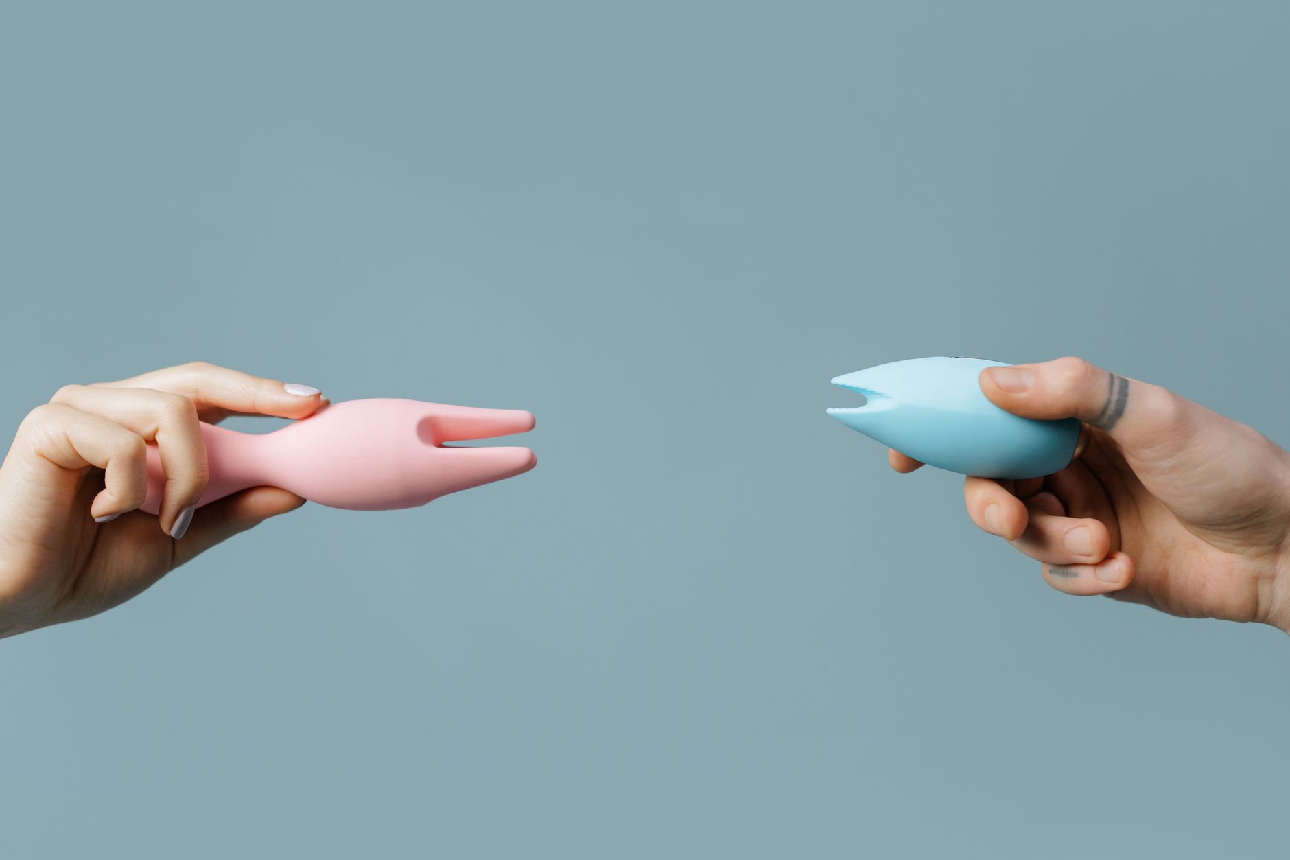 Twee handen houden roze en blauwe siliconen vibrators vast tegen een pastelkleurige achtergrond.