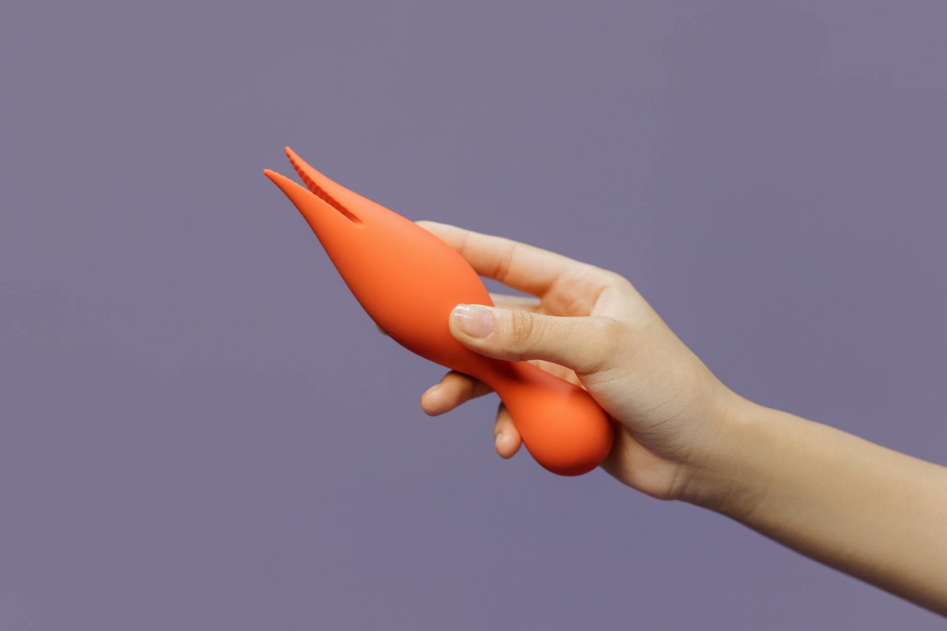 Een close-up van een hand met een oranje vibrator tegen een minimalistische paarse achtergrond, die moderne sensualiteit uitstraalt.